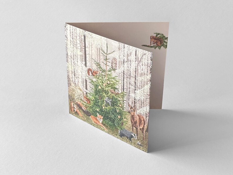 Op de afbeelding: Een gevouwen wenskaart met een bossc&egrave;ne met een versierde kerstboom. De illustratie bevat verschillende bosdieren zoals herten, vossen, eekhoorns en een das. De kaart heeft een lichtbeige binnenkant.