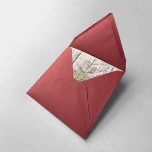 Op de afbeelding: Een rode envelop met een gedeeltelijk zichtbare kaart erin. De kaart heeft een botanisch ontwerp met groene en bruine elementen. De envelop is open en toont het ontwerp van de kaart.