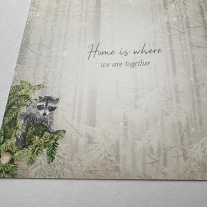 Op de afbeelding: Een wenskaart met een aquarel illustratie van een wasbeer op een dennentak. De achtergrond toont een mistige bossc&egrave;ne met de tekst "Home is where we are together".