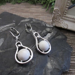 May include: A pair of silver-toned clip-on earrings. Each earring features a teardrop-shaped design with a white, spherical bead suspended within. The earrings are displayed on a dark, textured surface.