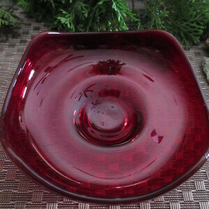 May include: A deep red glass dish with a rounded square shape and a raised center. The dish is made of clear glass with a deep red tint.