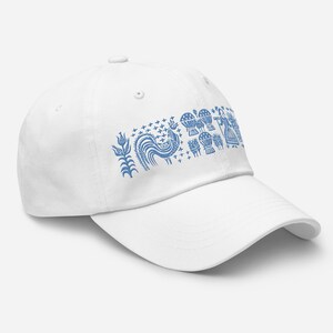 May include: White baseball cap with a blue folk art design. The design features a rooster, floral elements, and other stylized figures. The cap has a curved brim and an adjustable strap.