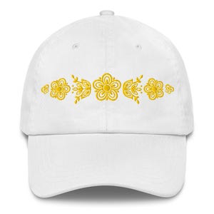 May include: White baseball cap with a curved brim. The cap features a gold floral embroidery design across the front. The embroidery includes flowers and leaves. The cap is made of a soft fabric.