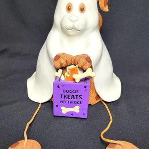 May include: A ceramic dog figurine with a white body, brown ears and paws, and a purple treat bag. The bag reads "Doggie Treats No Tricks" and contains a bone and a cat figurine. The dog has orange dangling feet.