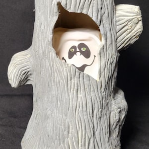 May include: A gray, tree-trunk-shaped Halloween decoration with a smiling ghost face peeking out from a hollowed-out section. The ghost has green eyes and a black mouth. The tree trunk has a textured, carved appearance.