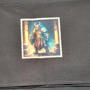 May include: Black fabric with a square image of a warrior in armor, holding a shield and sword. The warrior has a horned helmet and is standing in a stone structure. The image is centered on the fabric.