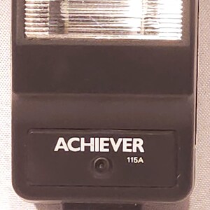 May include: A black Achiever 115A camera flash unit. The flash has a rectangular shape with a ridged light diffuser and a hot shoe mount. The word "ACHIEVER" is printed in white on the front.