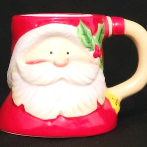 May include: A ceramic Christmas mug shaped like Santa Claus. The mug features a red base and rim, a white beard and face, and a cream-colored handle. Holly and berries decorate the side of the mug. The mug is a festive holiday decoration.