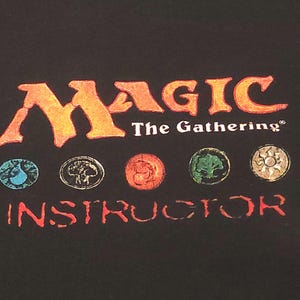 May include: Black fabric with the "Magic: The Gathering" logo in orange and white. Below the logo are five circular symbols in blue, gray, orange, green, and yellow. The word "INSTRUCTOR" is printed in orange below the symbols.
