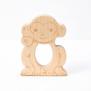 May include: A wooden monkey-shaped baby teether. The teether is made of light-colored wood and features engraved details of a monkey's face, body, and feet. The design includes a large opening in the center for easy gripping.
