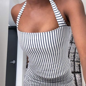May include: A white and navy blue striped halter top. The top features vertical stripes and a square neckline. The straps are also striped. The top is made of a lightweight fabric.
