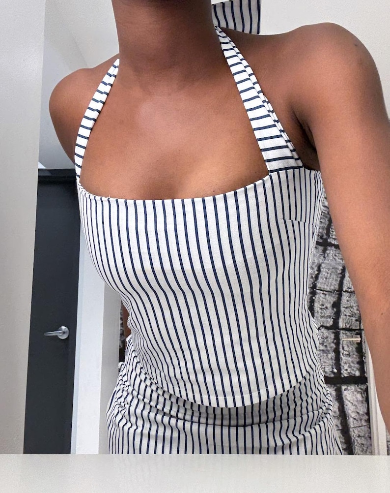May include: A white and navy blue striped halter top. The top features vertical stripes and a square neckline. The straps are also striped. The top is made of a lightweight fabric.