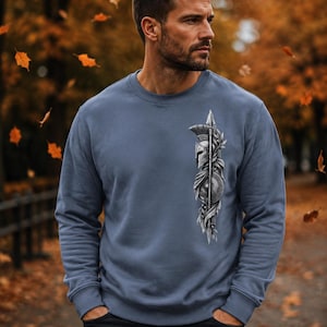 May include: A slate blue crewneck sweatshirt featuring a vertical graphic print on the left chest. The design depicts a detailed monochrome illustration of a Spartan helmet, a spear, and a round shield, all intertwined with decorative laurel leaves.