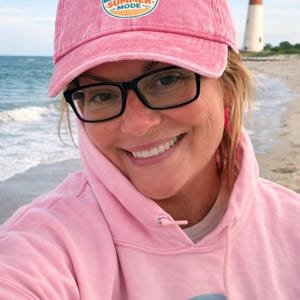 May include: A pink baseball cap with the text "SALTY SUMMER MODE" and a pink hoodie with the text "My cousin Bayes probed once". The image shows a person wearing glasses and smiling on a beach.