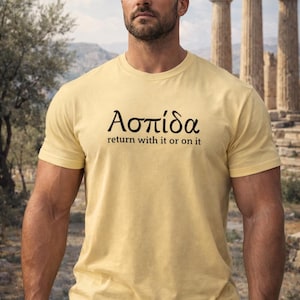 Può includere: Una maglietta giallo chiaro con la parola greca "Ασπίδα" e la frase "return with it or on it" stampate in nero. La maglietta è indossata da una persona davanti a uno sfondo architettonico classico.