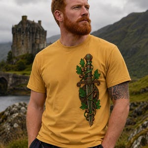 May include: A mustard yellow t-shirt featuring a detailed graphic print of a dagger entwined with green oak leaves and brown acorns. The dagger design includes intricate knotwork patterns along the hilt and blade, centered on the chest area.