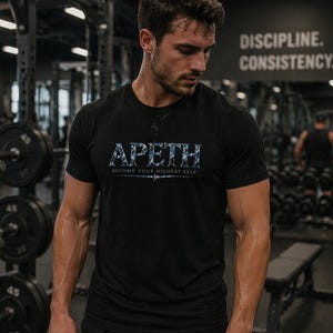May include: Black crew neck t-shirt with the word "APETH" in a blue and white floral pattern across the chest. Below the word is the phrase "BECOME YOUR HIGHEST SELF". The image is taken in a gym setting.