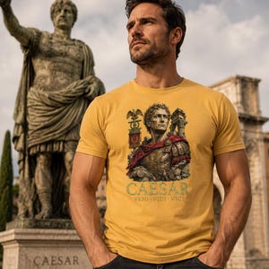 May include: A mustard yellow short-sleeved t-shirt featuring a graphic print of Julius Caesar in armor with a red cloak, a Roman standard, and architectural ruins. The text CAESAR VENI VIDI VICI is printed in a light green font below the illustration.