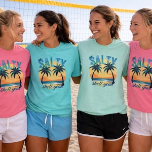 Puede incluir: Cuatro mujeres con camisetas a juego con un diseño playero. Las camisetas son de color rosa y turquesa, con palmeras, una puesta de sol y las palabras "Salty Shell Yeah". Están de pie en una playa de arena.