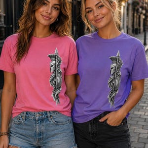 May include: A pink t-shirt and a purple t-shirt, each featuring a graphic print on the left chest. The print depicts a detailed grayscale illustration of a Spartan helmet, a spear, and a shield, surrounded by stylized laurel leaf accents.