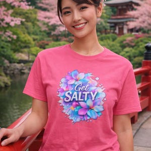 Puede incluir: Una camiseta rosa con el texto "Get Salty" rodeado de un diseño floral en tonos azules, morados y blancos. La camiseta de manga corta es usada por una persona en un entorno al aire libre.