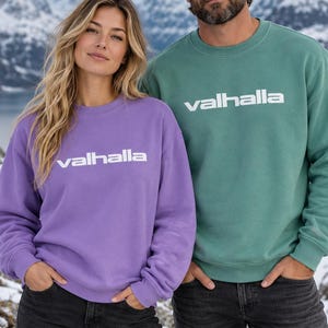 May include: A purple crewneck sweatshirt and a green crewneck sweatshirt, both featuring the word valhalla printed in white across the chest. The sweatshirts have long sleeves, ribbed cuffs, and a ribbed neckline, paired with dark grey denim jeans.