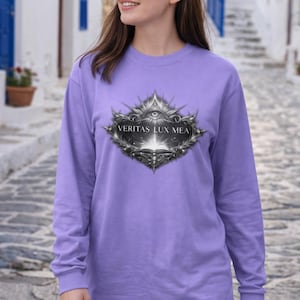 May include: A long-sleeved lavender shirt featuring a detailed silver emblem with the text "VERITAS LUX MEA." The emblem has an eye symbol and ornate, spiky edges. The shirt is made of a soft material.