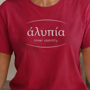 Può includere: T-shirt bordeaux con testo bianco curvo. Il testo recita "άλυπία" sopra "Inner stability". La maglietta è a maniche corte e realizzata in materiale morbido.
