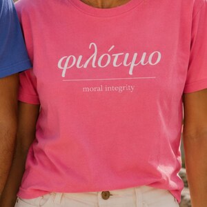 Puede incluir: Una camiseta rosa brillante con la palabra griega "φιλότιμο" y la traducción al inglés "moral integrity" impresa en blanco. La camiseta se combina con pantalones cortos blancos. El estilo general es casual y veraniego.