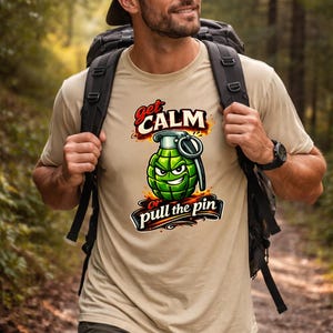 May include: Tan t-shirt with a graphic of a green cartoon grenade with the text "Get Calm or Pull the Pin". The shirt is worn by a person wearing a backpack and a cap.