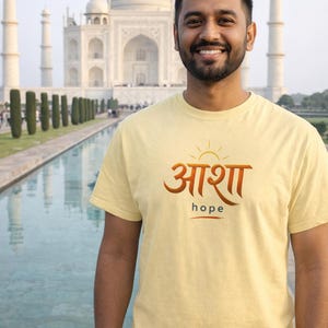 Może przedstawiać: Uśmiechnięta osoba w jasnożółtej koszulce z napisem "आशा" i angielskim słowem "hope". W tle Taj Mahal.