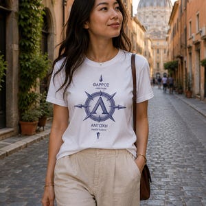 May include: A white short-sleeved t-shirt featuring a blue graphic of a compass-like design with a central Greek letter Lambda. The text reads ΘΑΡΡΟΣ courage and ΑΝΤΟΧΗ endurance. The shirt is paired with beige trousers and a brown shoulder bag.