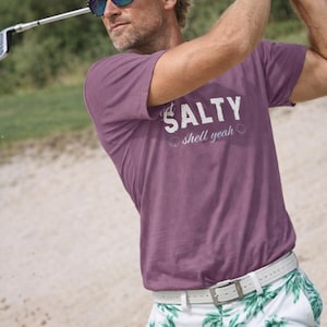 Può includere: Una persona che gioca a golf, indossa una maglietta viola con la scritta "get salty shell yeah", occhiali da sole e pantaloncini bianchi con stampa di palme. La persona indossa una cintura bianca e un guanto da golf.
