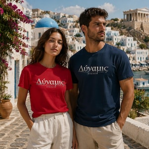Könnte beinhalten: Ein Mann und eine Frau tragen passende T-Shirts mit griechischem Text. Das Shirt der Frau ist rot, das des Mannes marineblau. Der Text auf beiden Shirts lautet "Δύναμις" über "Strength" in Silber. Sie stehen vor einer Küstenstadt.
