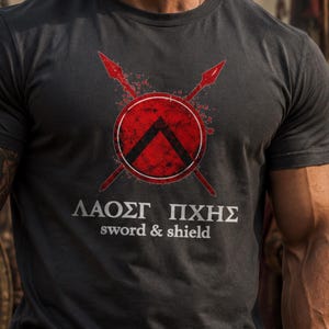 Op de afbeelding: Een antracietgrijs T-shirt met ronde hals en een afbeelding van twee gekruiste rode speren achter een rood rond schild met een zwart lambda-symbool. Onder de afbeelding staat de tekst "ΛΑΟΣΓ ΠΧΗΣ" in witte hoofdletters en "sword & shield".
