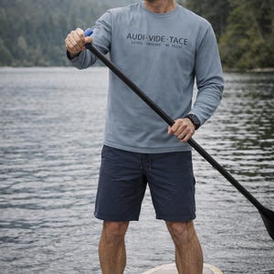 May include: A person on a paddleboard wearing a long-sleeved, light blue shirt with the text "AUDI VIDE TACE" and dark blue shorts. The person is holding a paddle with a black shaft and blue handle. The background shows a lake and mountains.