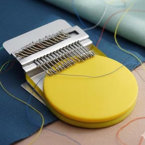 May include: A yellow thread organizer with a silver metal comb-like structure. The organizer is designed to hold multiple threads, with a circular base and a rectangular top. Threads of various colours are visible.