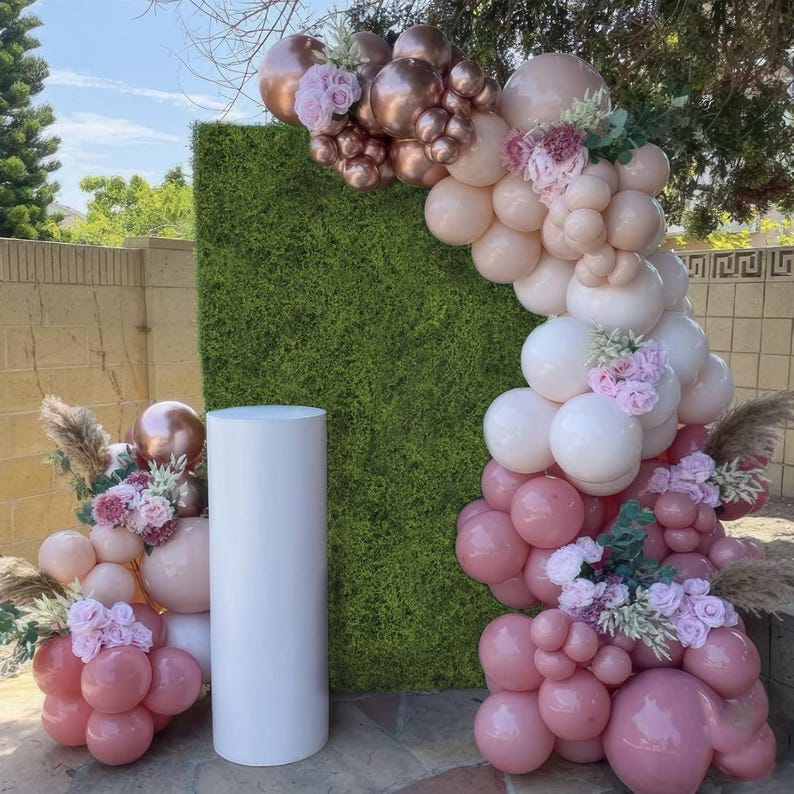 May include: A green artificial grass backdrop panel with a white cylindrical pedestal and a balloon garland featuring rose gold, blush pink, and cream balloons, accented with artificial pink roses and dried pampas grass plumes.