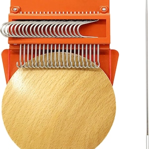 May include: An orange and wood-toned weaving tool with metal hooks and a wooden base. A long, thin needle is positioned to the right. The tool is designed for creating woven crafts and textiles.