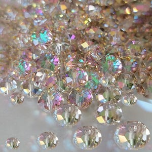 May include: A collection of iridescent crystal beads in various sizes. The beads have multiple facets that reflect light, creating a rainbow effect. The beads are clear with hints of pink, green, and yellow.