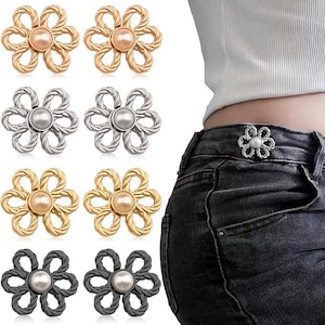 May include: A set of eight flower-shaped waist tightening pins in gold, silver, and black tones, each featuring a central faux pearl. The pins have a twisted metal wire design forming six petals and are shown attached to the waistband of black jeans.