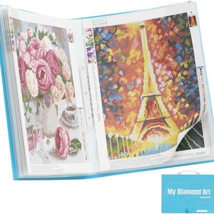 May include: A blue diamond art storage book with two completed diamond paintings visible. One painting depicts a vase of pink and white flowers, and the other shows the Eiffel Tower. The book has a handle and the text "My Diamond Art".