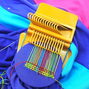 May include: A yellow plastic weaving loom with a partially woven circular piece of blue fabric. The weaving uses colorful threads of red, yellow, green, and blue. The loom is set against a backdrop of blue, pink, and teal fabric.