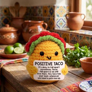 May include: A yellow crocheted taco plush toy with black eyes and a small smile. It features a red and green topping and holds a white sign that reads: POSITIVE TACO. It's okay to fall apart sometimes. Tacos fall apart & we still love them. Tiny taco has your back whenever you need a pick-me-up.