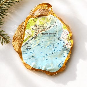 May include: An oyster shell with a gold-painted exterior and a decoupage map of Higgins Beach on the interior. The map features light blue water, green land areas, and black text labels including Higgins Beach and Cool Rock with depth numbers.