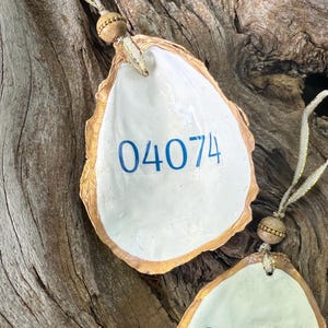 May include: Two oyster shell ornaments with gold trim and the number "04074" in blue. Each shell is attached to a gold ribbon with a wooden bead. The shells are displayed on a piece of driftwood.