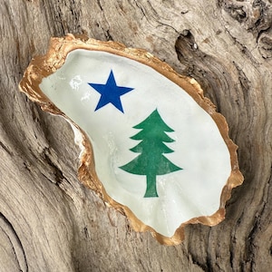 May include: An oyster shell dish with a gold-painted rim. The interior is painted white with a blue star and a green pine tree design. The dish is displayed on a piece of weathered wood.