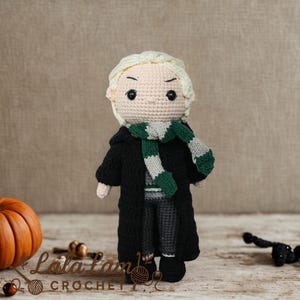 Puede incluir: Muñeca de ganchillo hecha a mano de un personaje con cabello rubio, que lleva un abrigo negro, pantalones grises y una bufanda verde y blanca. Dos calabazas naranjas y el texto "Lola Lamb Crochet" también están en la imagen.