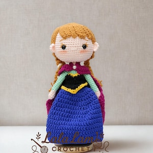 Puede incluir: Mu&ntilde;eca de ganchillo hecha a mano de un personaje con cabello casta&ntilde;o rojizo en trenzas, que lleva un vestido azul, una blusa negra con ribetes amarillos y una capa morada. La mu&ntilde;eca tiene un tono de piel claro y est&aacute; sobre una base de madera.