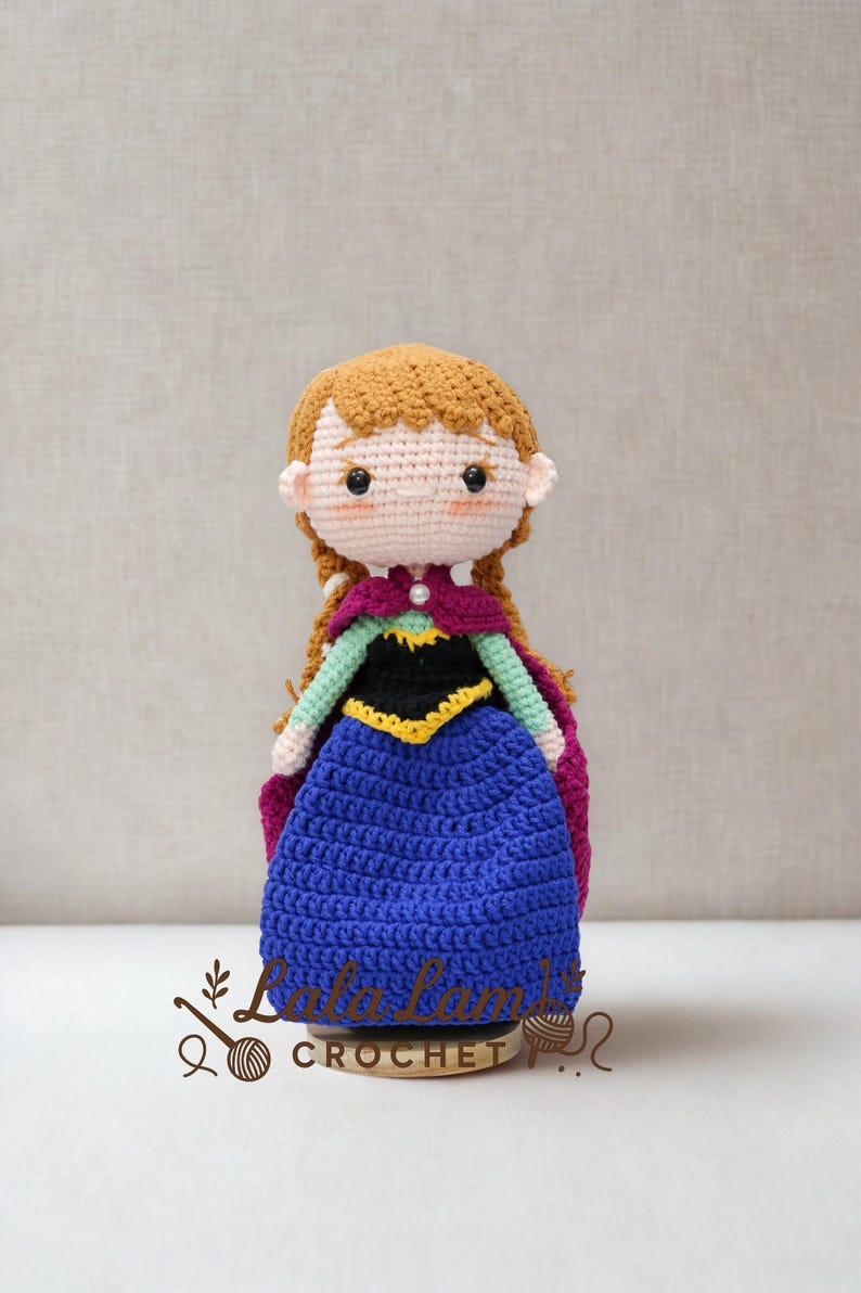 Puede incluir: Mu&ntilde;eca de ganchillo hecha a mano de un personaje con cabello casta&ntilde;o rojizo en trenzas, que lleva un vestido azul, una blusa negra con ribetes amarillos y una capa morada. La mu&ntilde;eca tiene un tono de piel claro y est&aacute; sobre una base de madera.